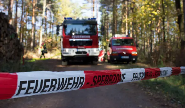 Symboolfoto van twee Duitse brandweerwagens in een bosgebied met een afzetlint ervoor waarop Feuerwehr - Sperrzone staat.