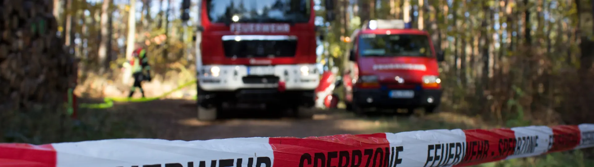 Symboolfoto van twee Duitse brandweerwagens in een bosgebied met een afzetlint ervoor waarop Feuerwehr - Sperrzone staat.